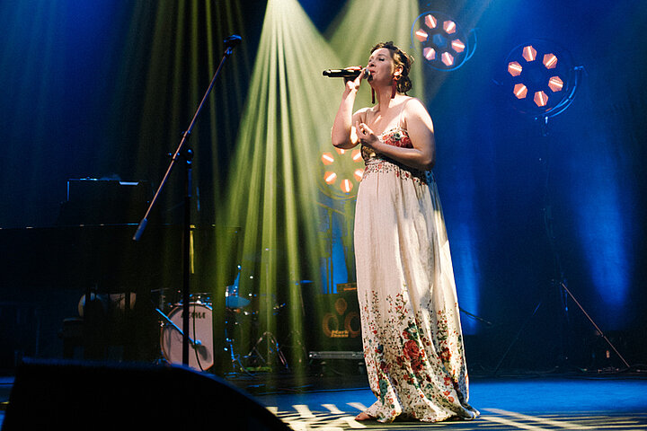 Zdjęcie piosenkarki występującej na scenie. Ma na sobie jasną sukienkę w ozdobioną haftowanymi kwiatami.