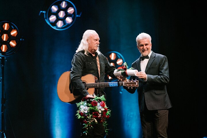 Zdjęcie przedstawiadwóch mężczyzn na scenie. Jeden z nich trzyma gitarę. Drugi wręcza mu dyplom.